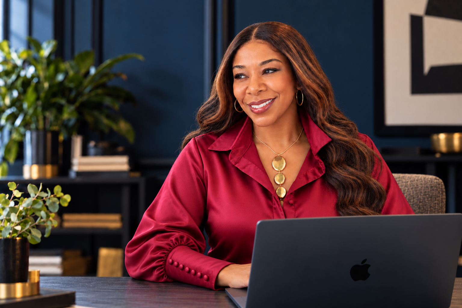 Ursula Spencer in burgundy satin, seated confidently behind a black MacBook in a navy editorial workspace